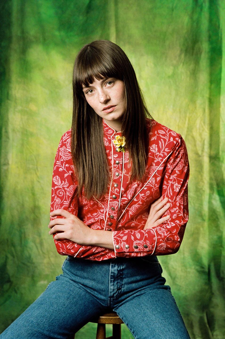 A woman with long hair and bangs sits on a stool against a green backdrop, wearing a red floral shirt and jeans.