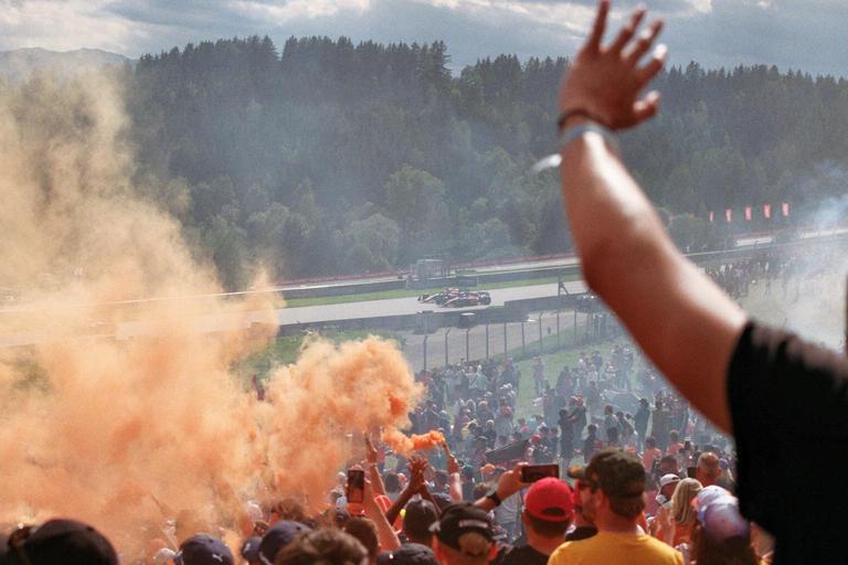 A crowd of spectators watches a car race from a hillside, with orange smoke rising from the crowd.