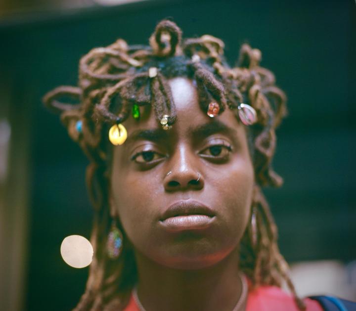 A woman with dreadlocks adorned with beads and shells looks directly at the viewer.