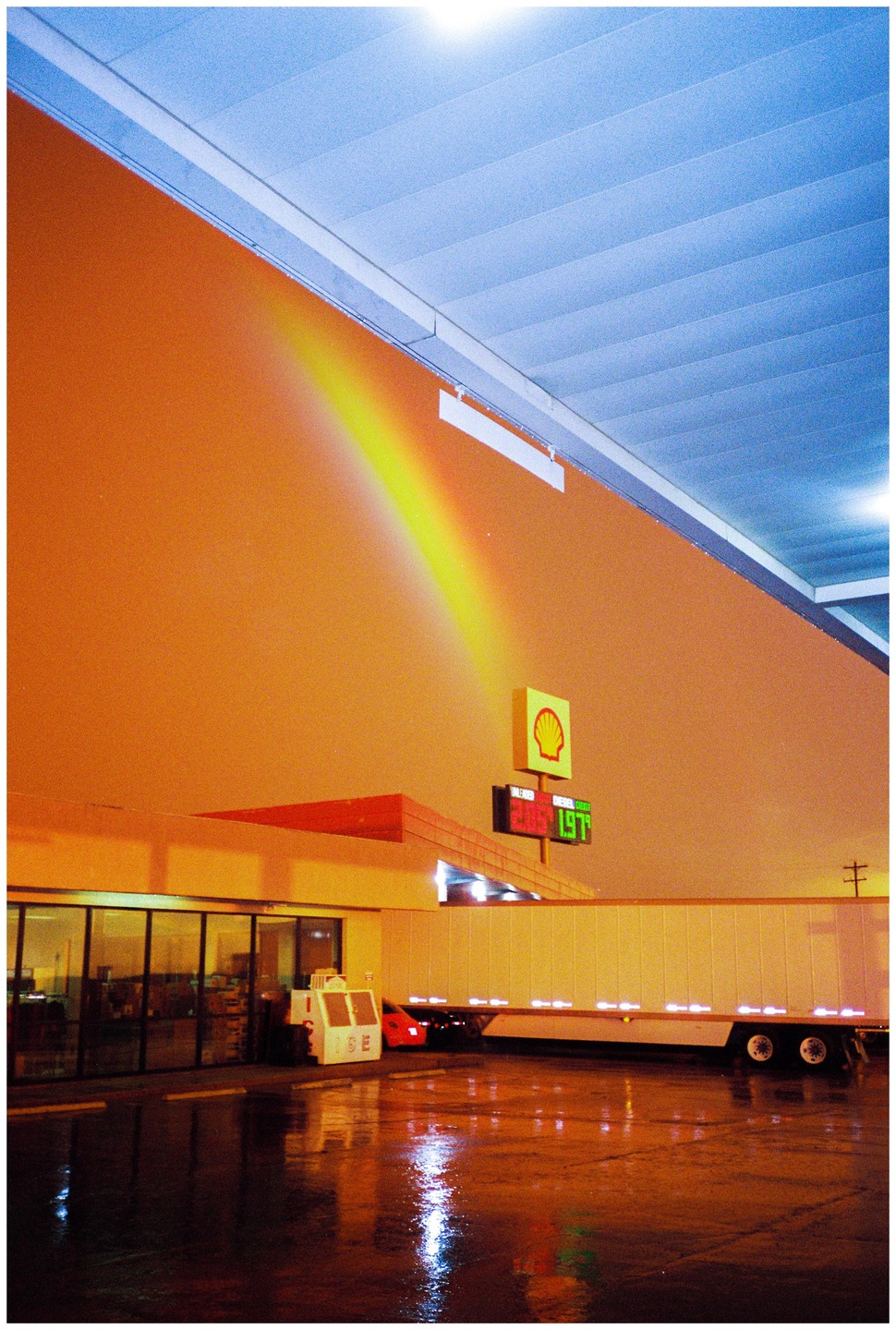 A rainbow-like light streak appears on the side of a building at a gas station at night, with a semi-trailer truck parked nearby.