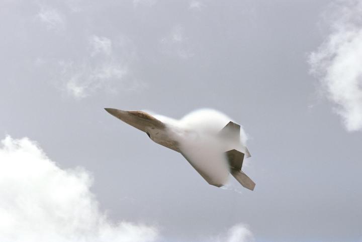 A jet aircraft is flying through the sky with a vapor cone forming around its body.