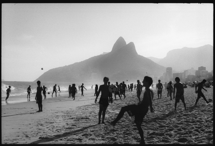 A group of people play soccer on a beach with mountains and a city in the background.