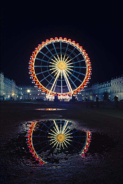 A brightly lit ferris wheel is reflected in a puddle on the ground at night.
