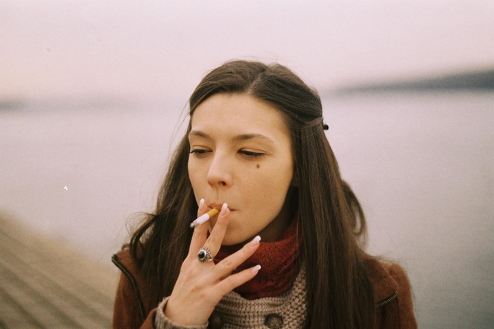 A woman with long brown hair and a brown jacket smokes a cigarette while looking down.