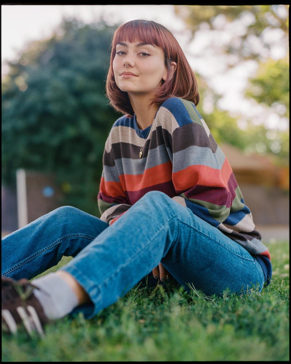 A woman with red hair and a striped sweater sits on the grass.
