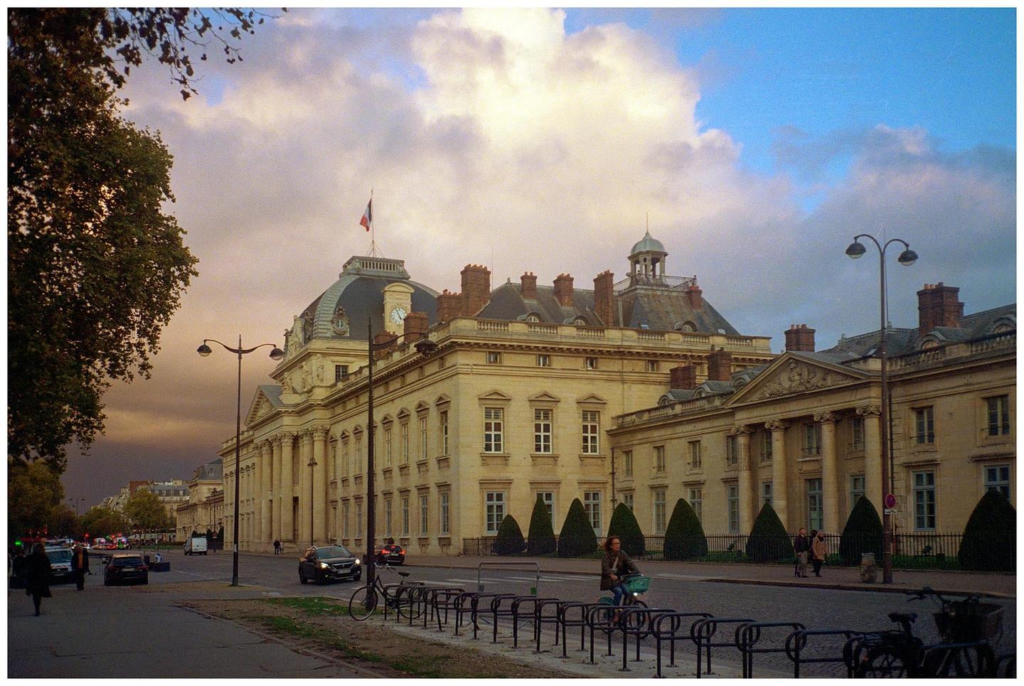 A large classical building with a domed roof and a French flag on top stands next to a street with cars and people.