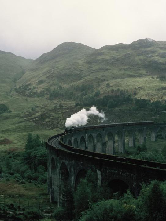 A steam train travels across a long, arched viaduct through a mountainous, green landscape.