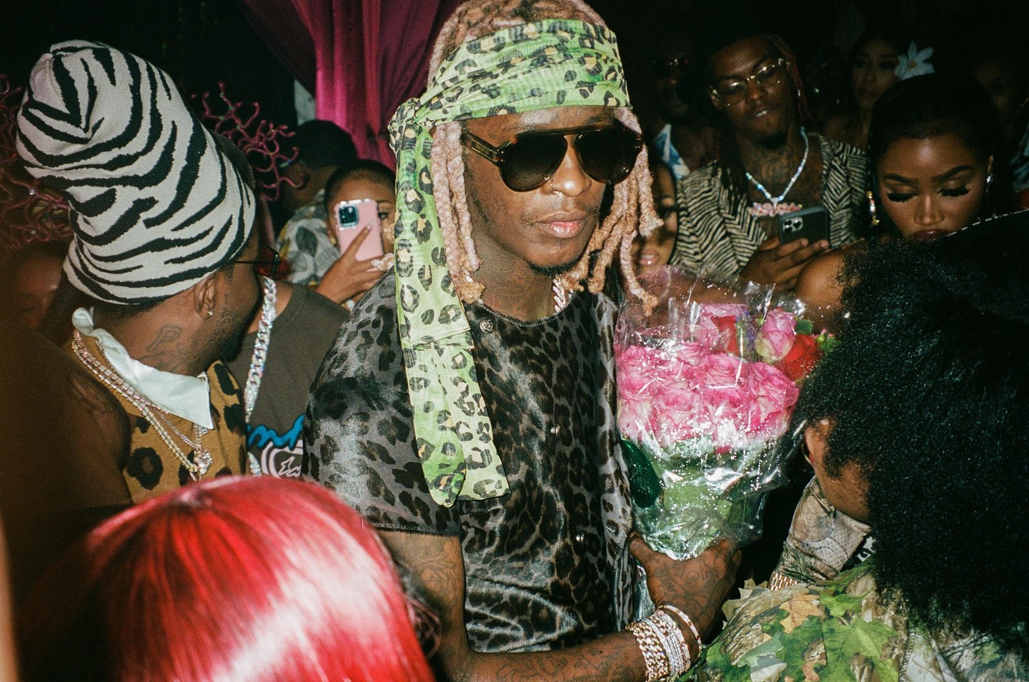 A man in sunglasses and a leopard print shirt holds a bouquet of pink roses at a party.