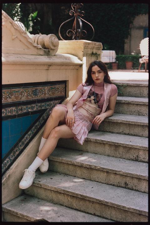 A woman wearing a pink outfit with a cat graphic on her shirt sits on stone steps next to a tiled wall.