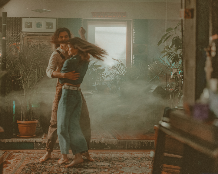 A couple dances barefoot on a patterned rug in a room filled with plants and haze.