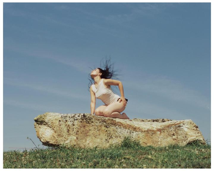 A woman kneels on a large rock with her head tilted back and hair blowing in the wind under a clear sky.