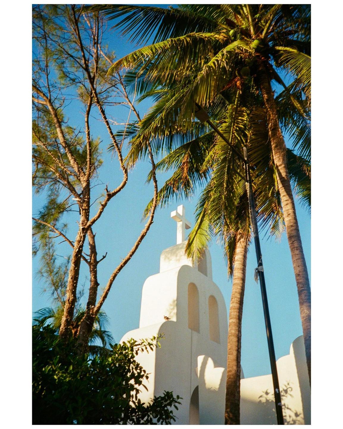 A white church with a cross on top is framed by palm trees and other foliage against a clear blue sky.