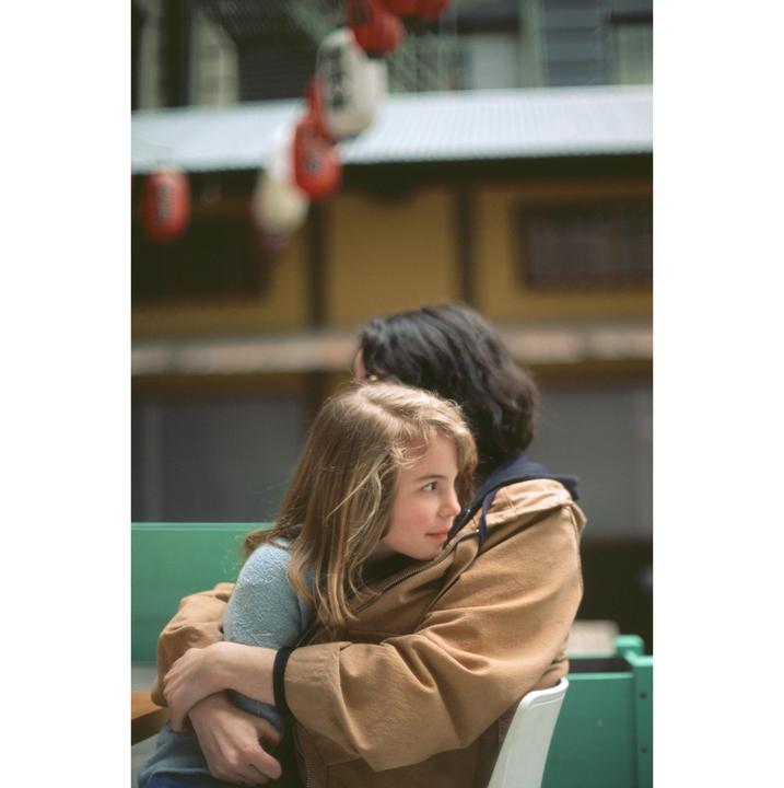 A girl with long blond hair is embraced by an adult with dark hair, sitting on a green bench with red and white lanterns in the background.