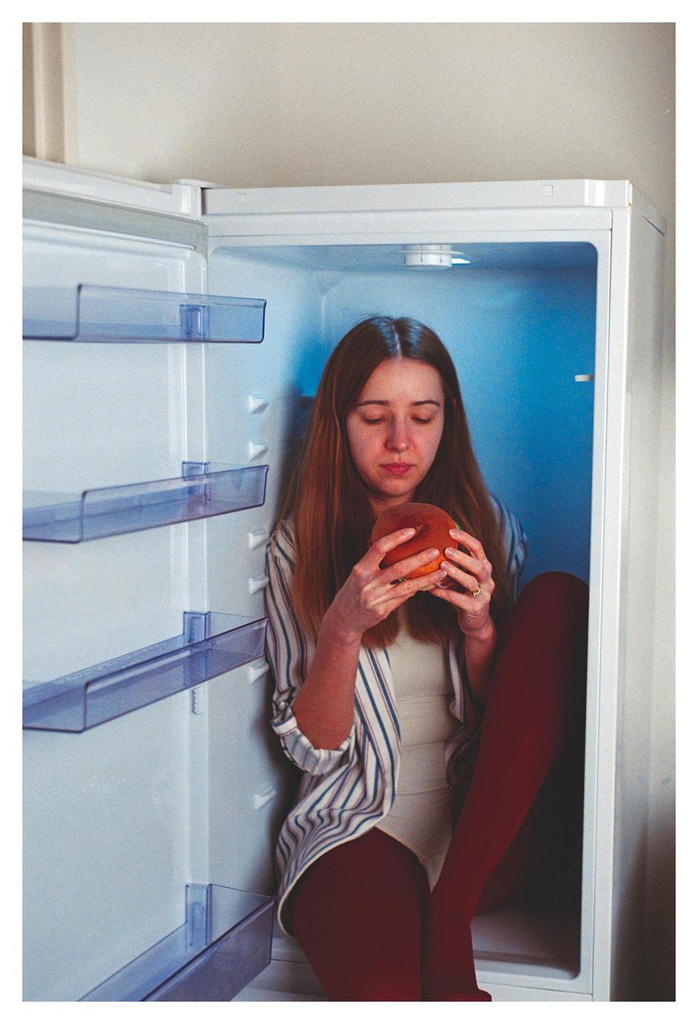 A woman sits inside an empty refrigerator, holding a peach.