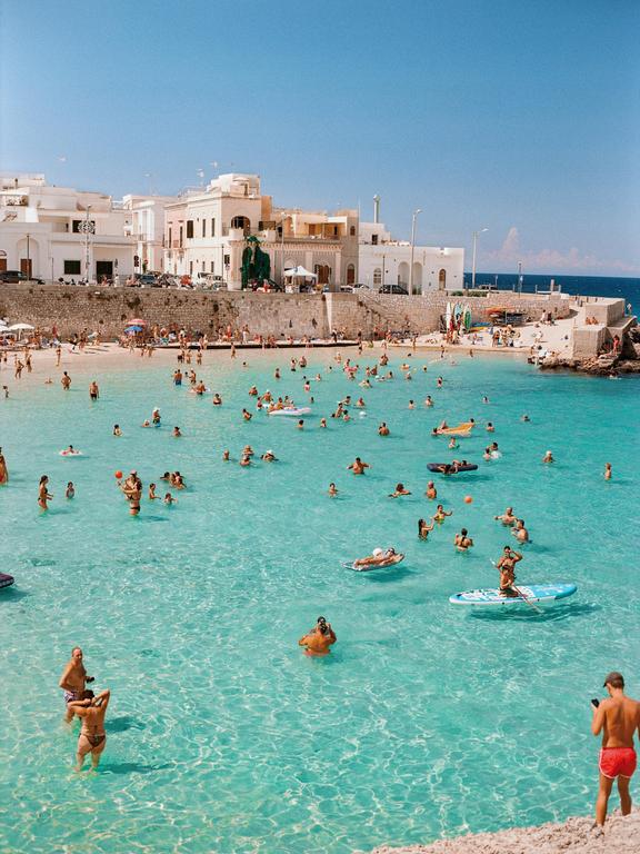 Many people swim in the clear blue water of a beach next to a town with white buildings.