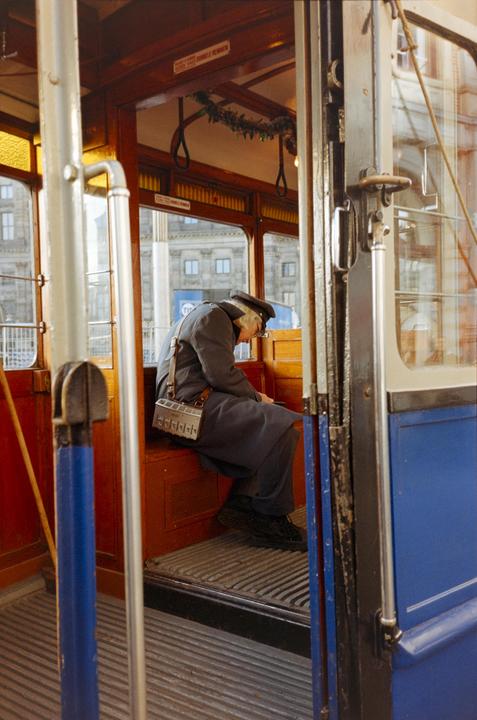 A man in uniform sits inside a tram with a ticket dispenser hanging from his shoulder.