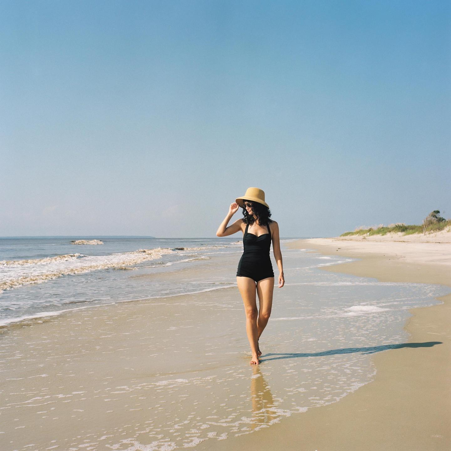 A woman in a black swimsuit and straw hat walks along a sandy beach with waves gently breaking on the shore.
