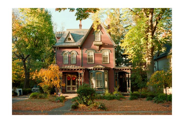 A large, ornate house with a red exterior and green trim is surrounded by trees with autumn foliage.