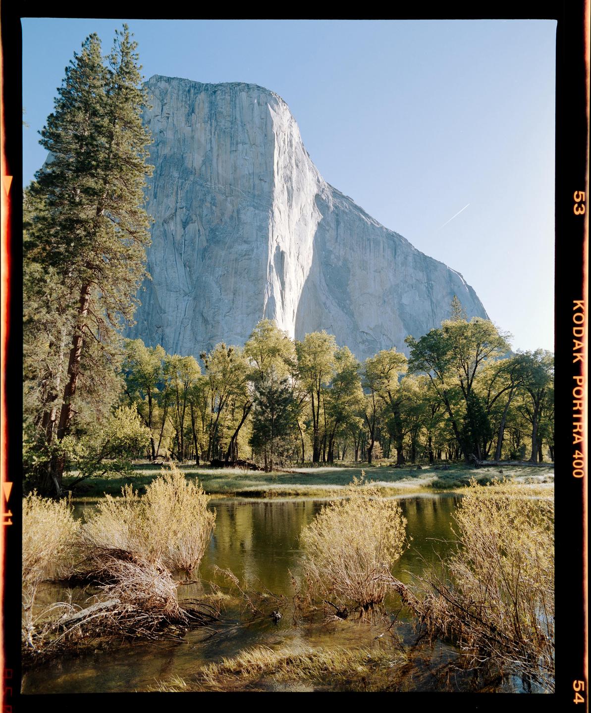 A large mountain cliff rises above a body of water surrounded by trees and brush.