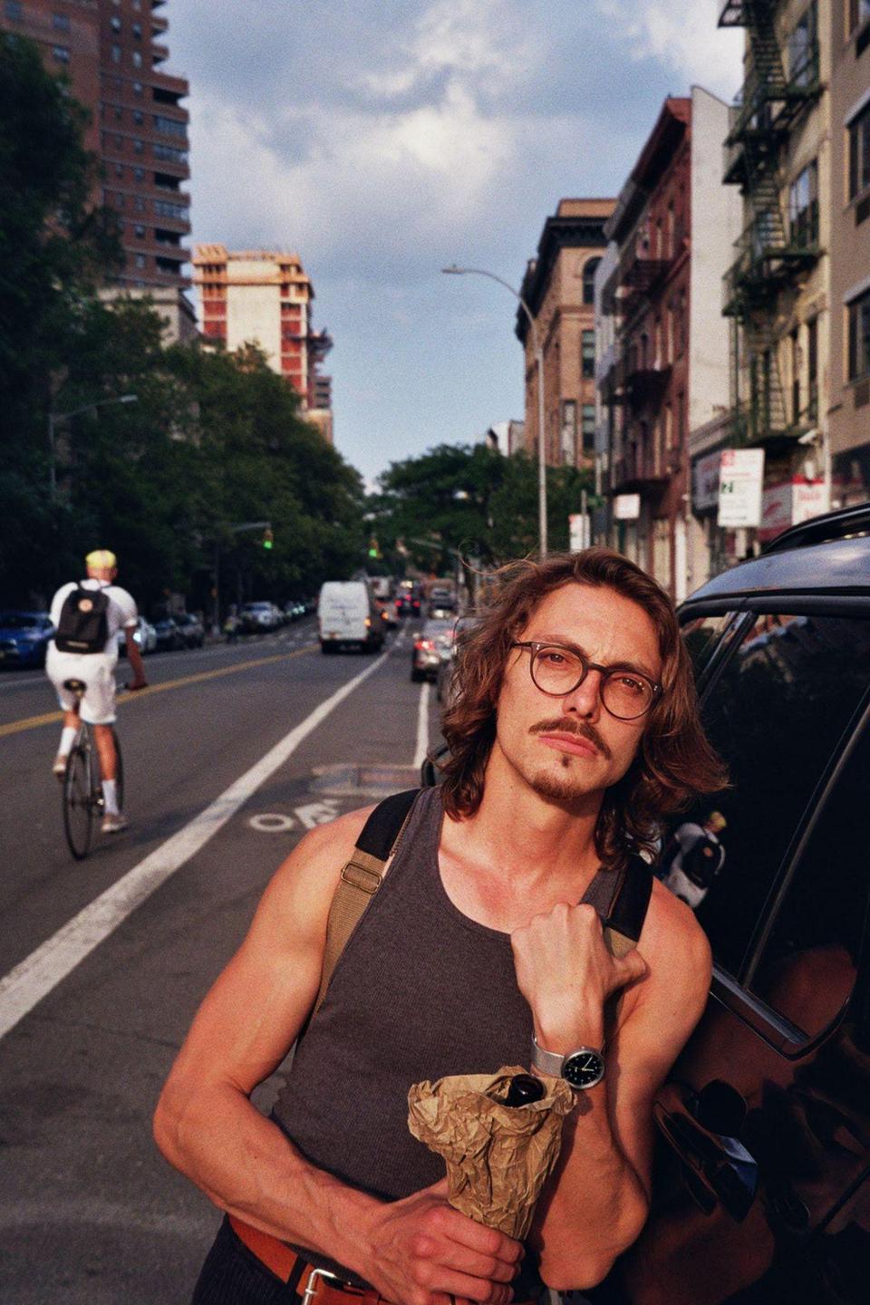 A man with a mustache and glasses stands on a city street holding a paper bag, with a cyclist and buildings in the background.