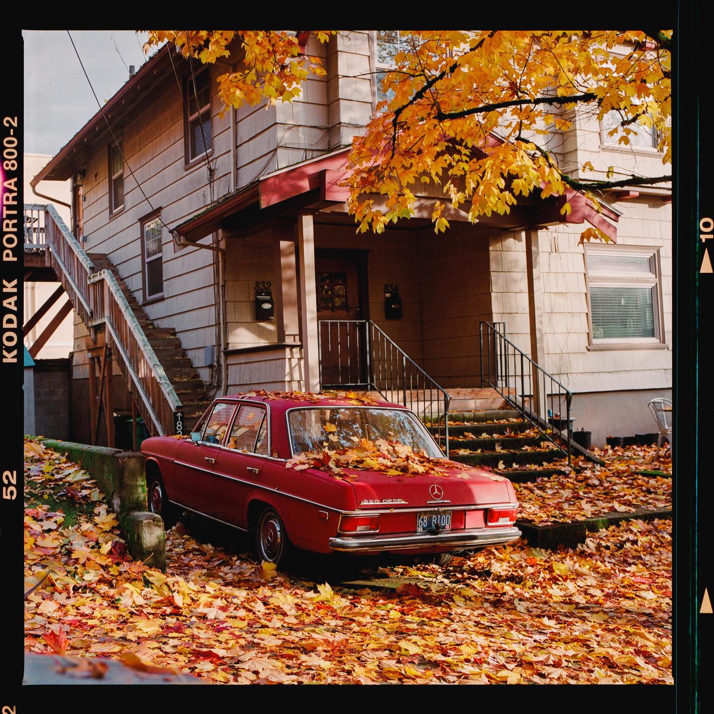 A red car is parked in front of a house, surrounded by autumn leaves.