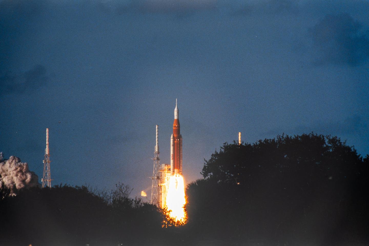 A rocket launches into a dark sky, with bright orange flames and smoke billowing from its base.