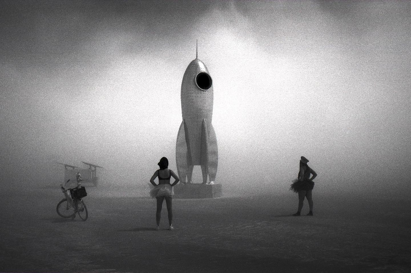 Two women stand in a hazy, open space with a large rocket sculpture and a bicycle.