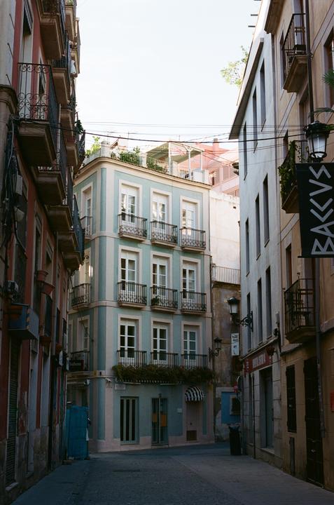 A narrow street is lined with tall buildings featuring balconies and a light blue corner building.