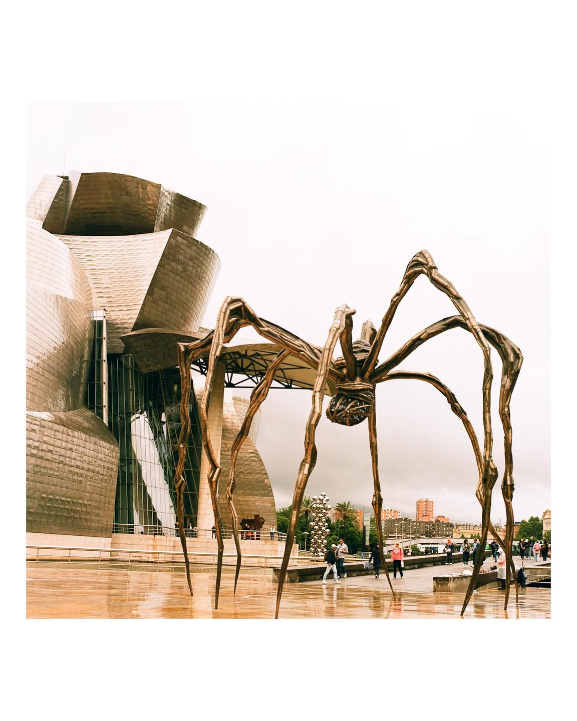 A large spider sculpture stands in front of a modern building with a reflective facade on a cloudy day.