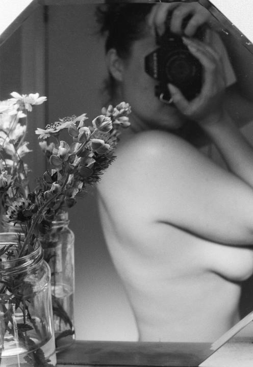 A woman takes a self-portrait in a mirror, with wildflowers in jars in the foreground.