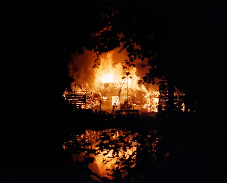 A house is engulfed in flames at night, with the fire reflecting on water in the foreground.