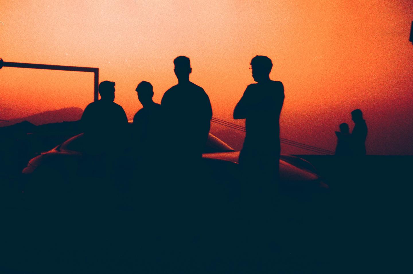 A group of men are silhouetted against a vibrant orange and red sky, with a car visible in the foreground.