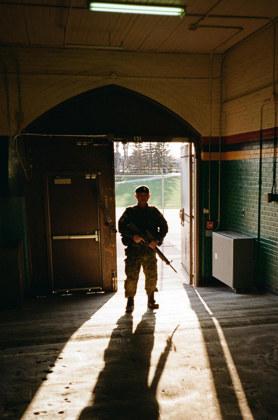 A man in military uniform stands in a doorway, holding a rifle, with strong backlighting creating long shadows on the floor.