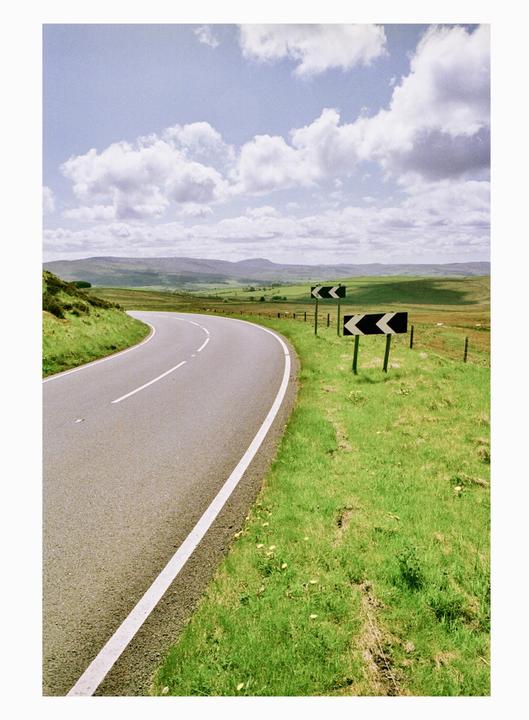 A winding road with two chevron signs on the right leads through a green, hilly landscape under a cloudy sky.