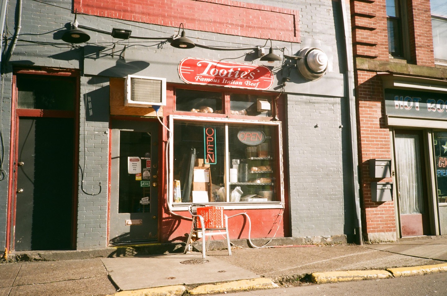 A red and gray brick building with a sign for "Tootie's Famous Italian Beef" has an open sign in the window and a red chair outside.