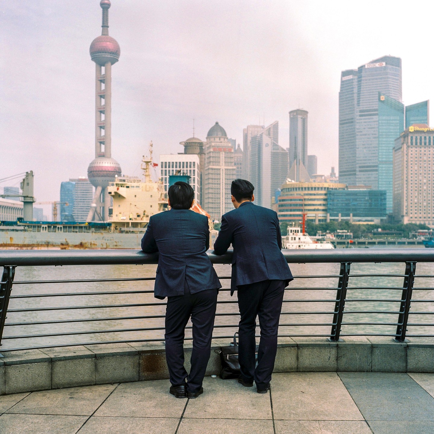 Two men in suits lean on a railing overlooking a river with a city skyline and a tower in the background.