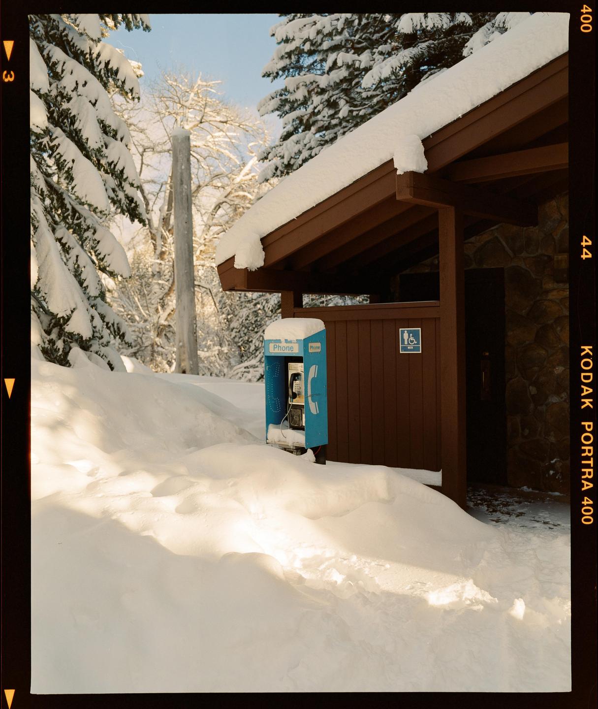 A blue payphone stands next to a brown building, surrounded by deep snow and snow-covered trees.