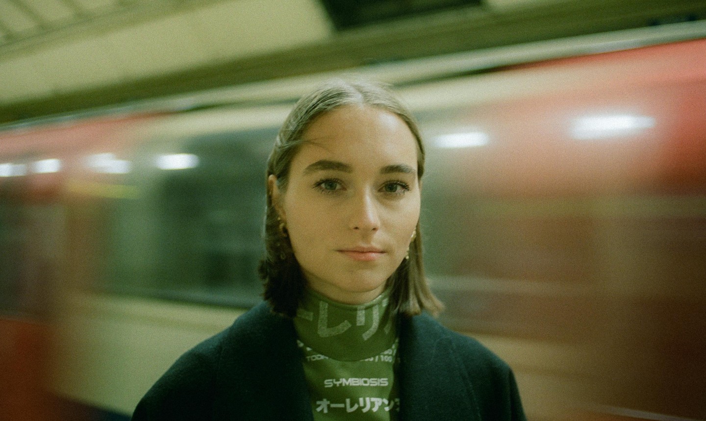 A woman stands on a subway platform with a blurred train passing behind her.