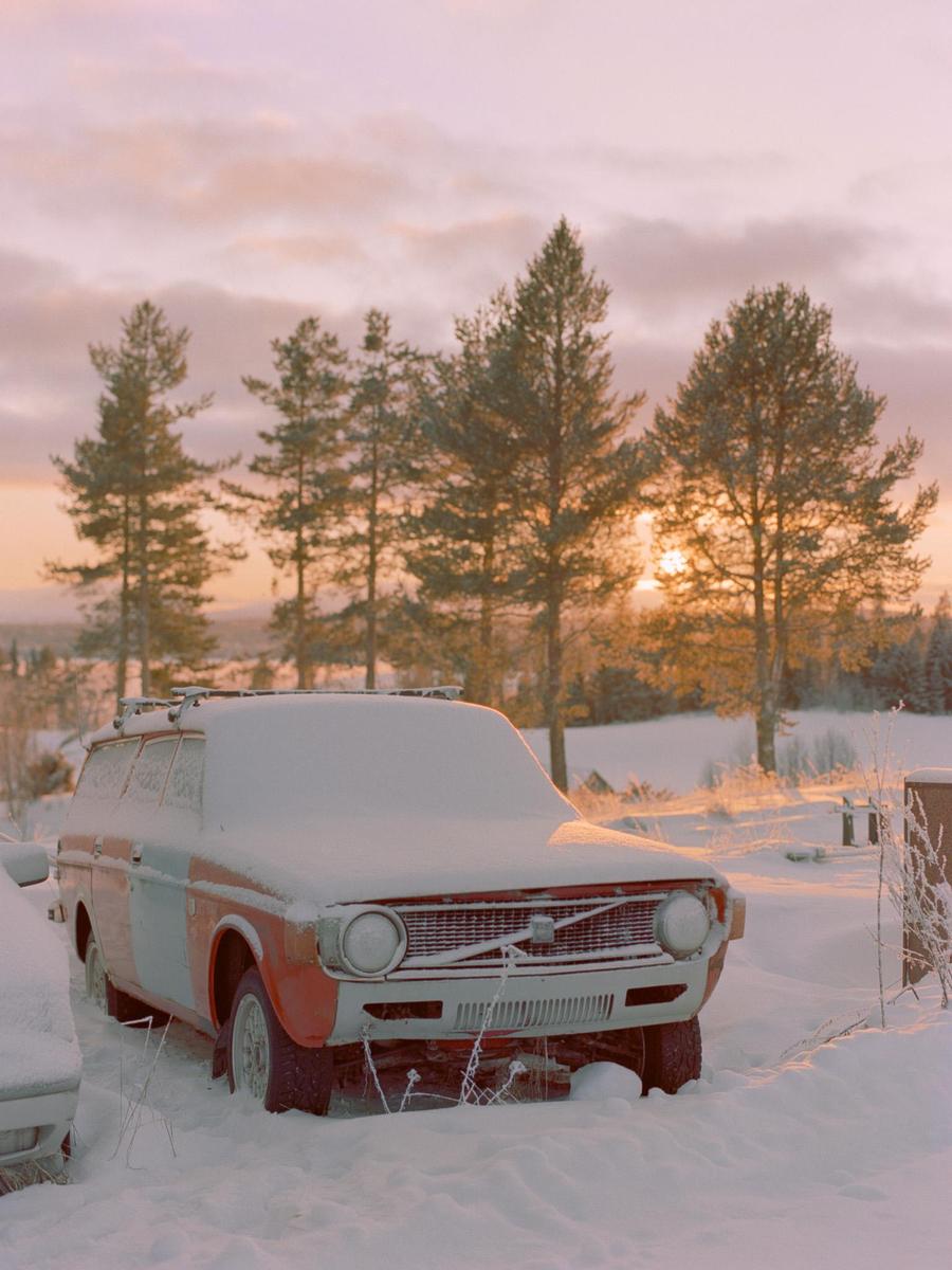 A snow-covered car is parked in a snowy landscape with pine trees and a sunset in the background.