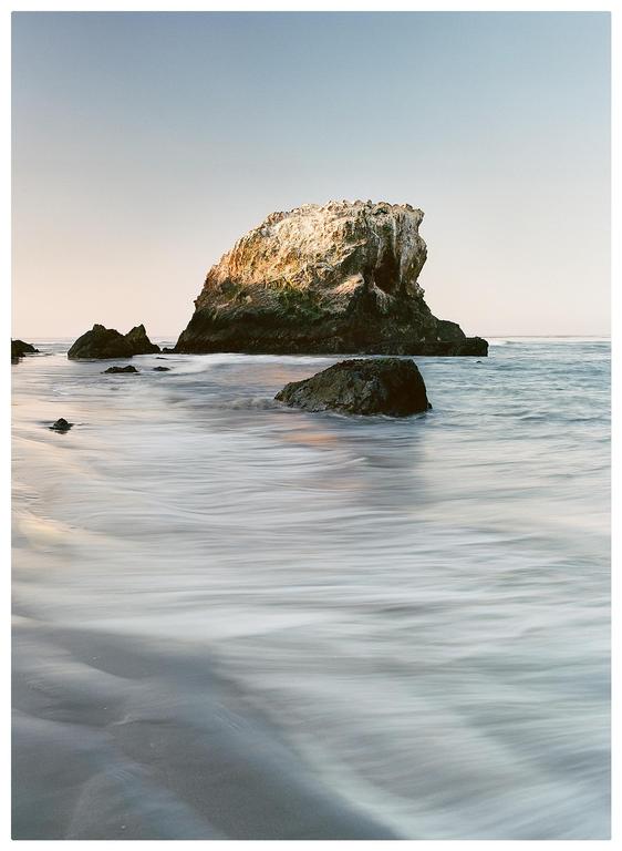The ocean washes over a sandy beach with several large rocks and a sea stack in the background.