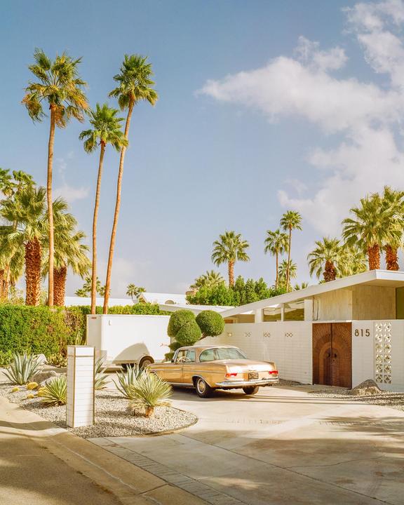 A gold car is parked in the driveway of a white house with palm trees and desert plants.
