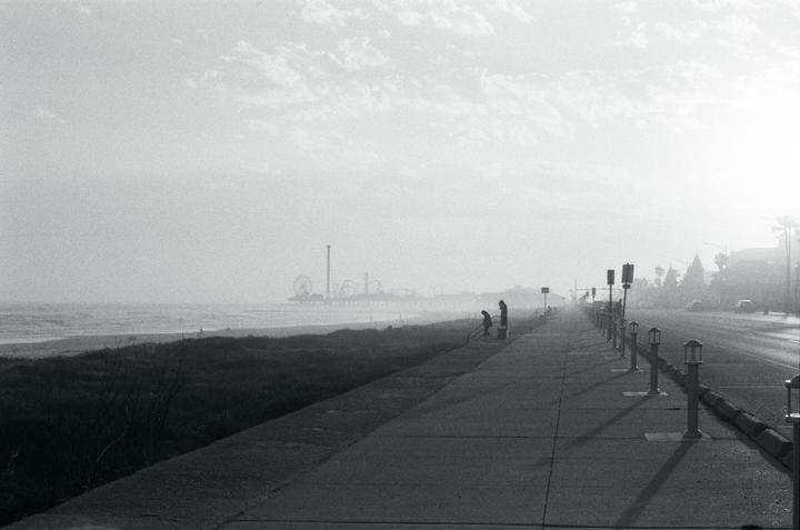 Two people stand on a boardwalk next to a beach with an amusement park in the distance.
