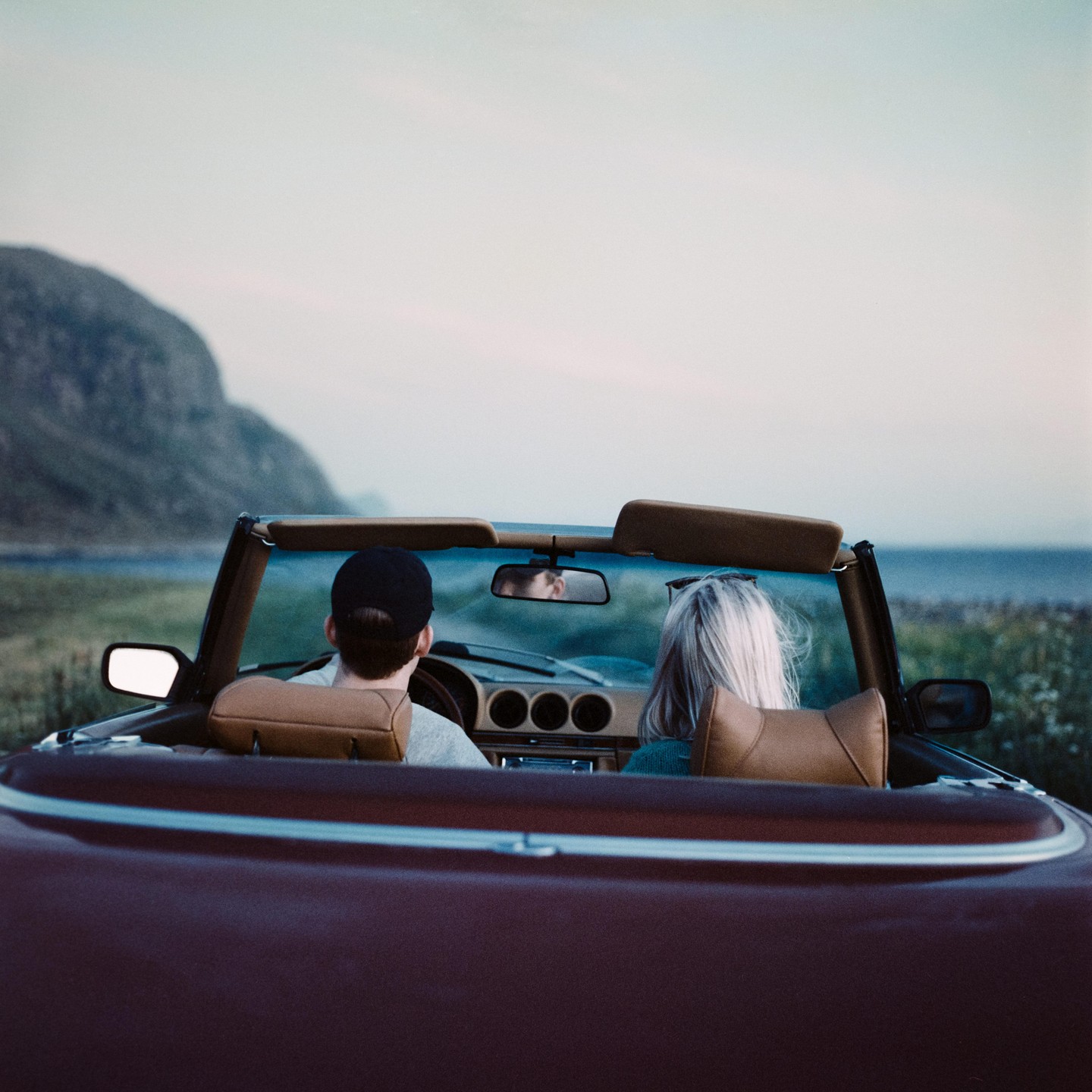 A couple drives a convertible car along a coastal road at sunset.
