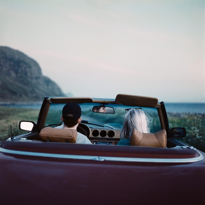 A couple drives a convertible car along a coastal road at sunset.
