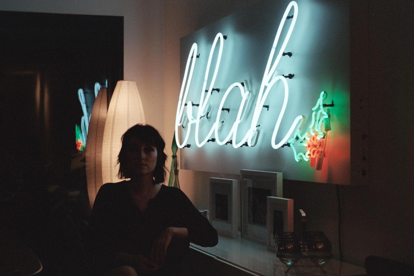 A woman sits in a dimly lit room with a neon sign that reads "blah" on the wall.