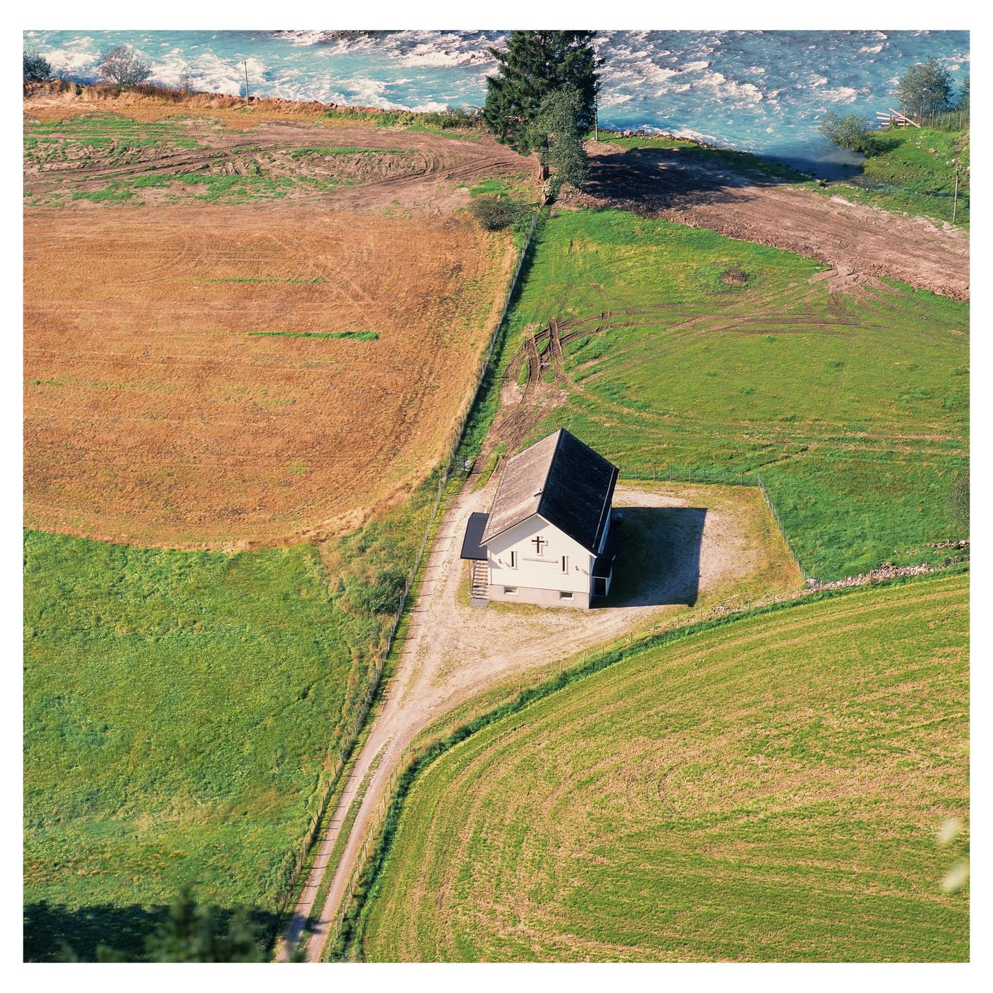 An aerial view shows a small house with a cross on its side, surrounded by fields and a river.