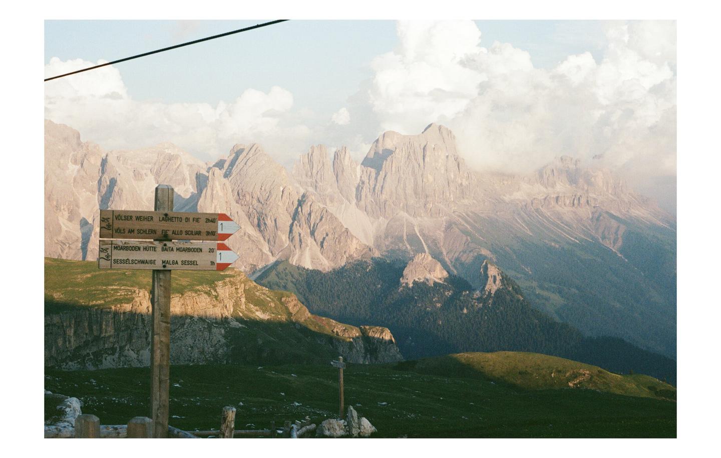 A wooden signpost with hiking directions stands in a grassy field with mountains and clouds in the background.