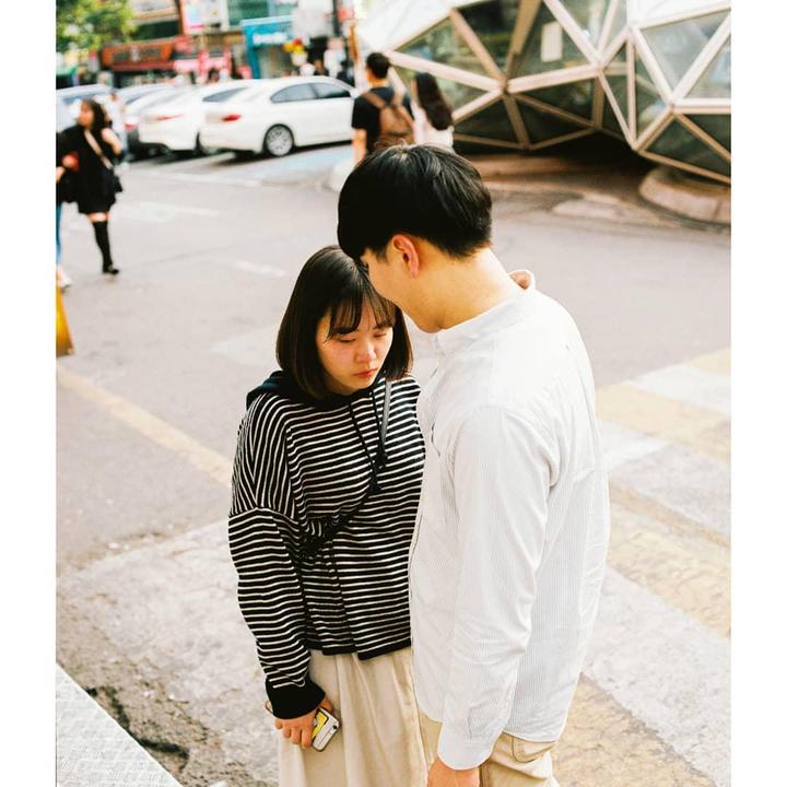 A man and a woman stand on a city street with a geometric building in the background.