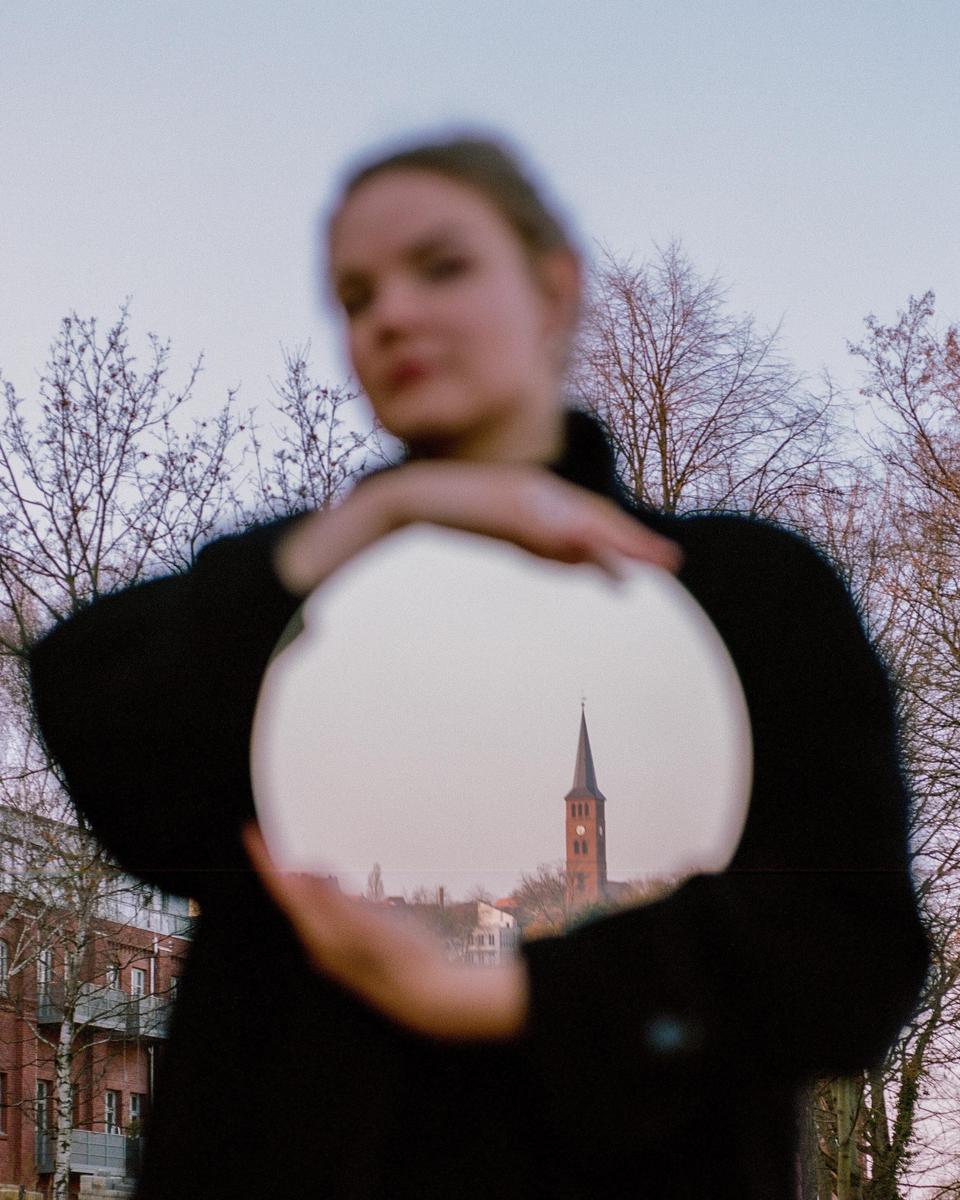A woman holds a mirror reflecting a church steeple in a city.