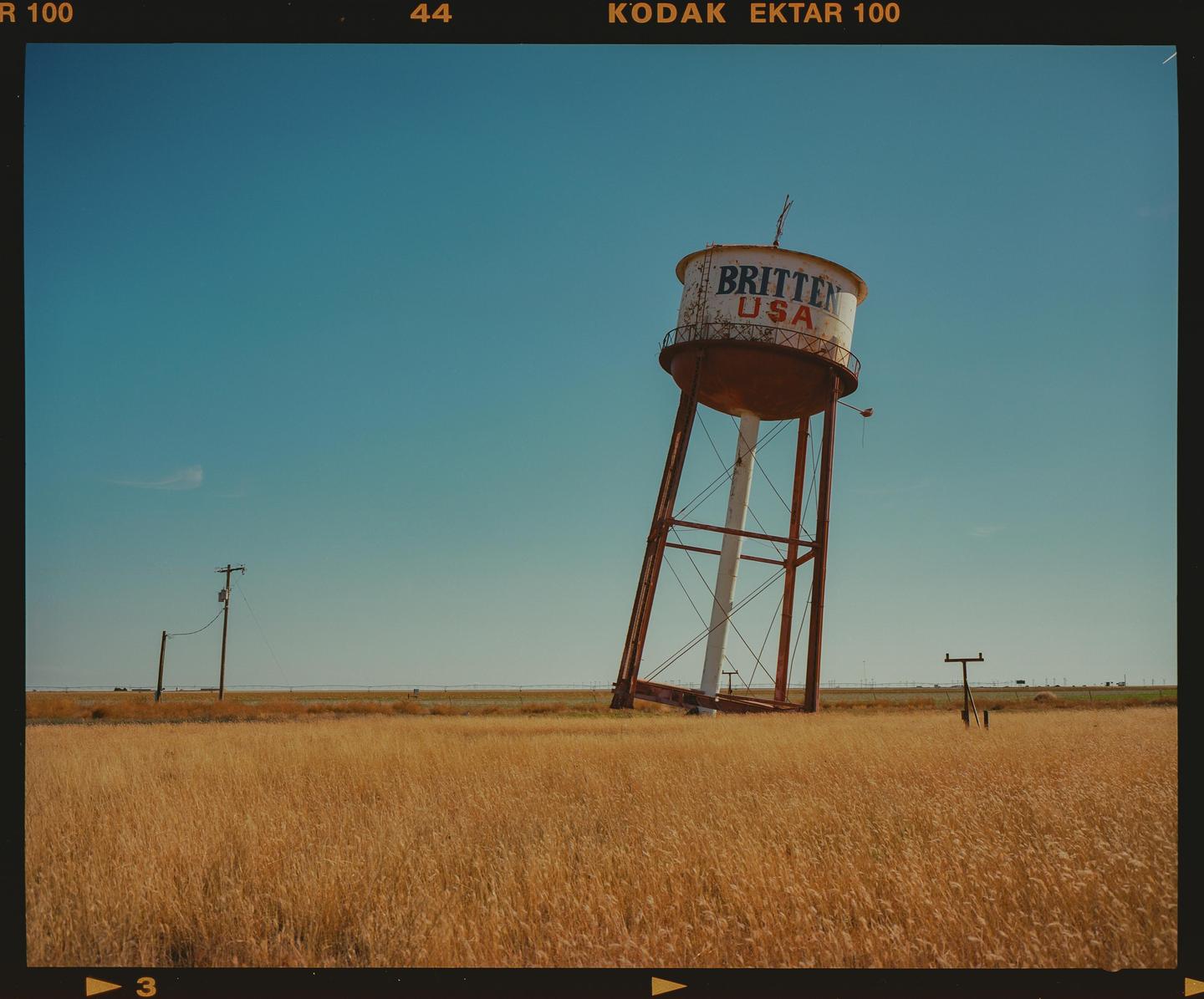 A leaning water tower with "BRITTEN USA" painted on it stands in a field of golden grass under a clear blue sky.